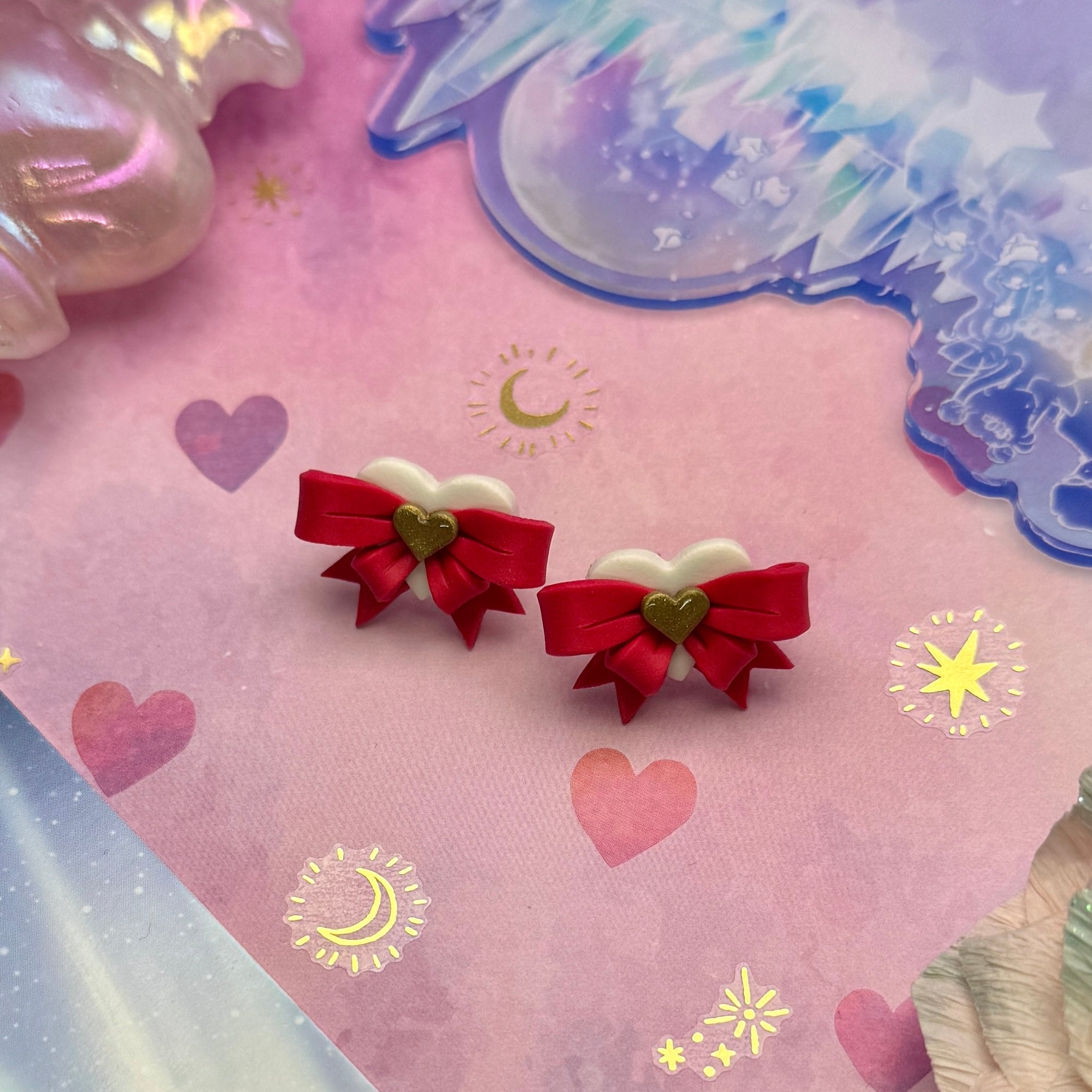 Pair of earrings with red bows and gold hearts on a decorative background with hearts and stars.