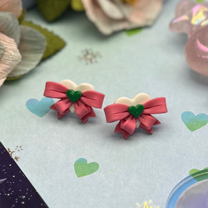 Pair of pink bow-shaped earrings with green centers on a light blue background with heart designs.
