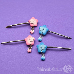 Four bobby pins with pink and blue polymer clay charms in the shape of sakura blossoms, displayed on a purple background.